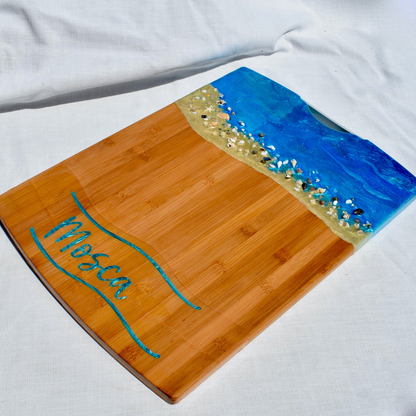 Beach themed charcuterie board features ocean shimmering ocean blue water, spiral and abalone shells, and is personalized with the buyers name between two to wavy teal colored lines.